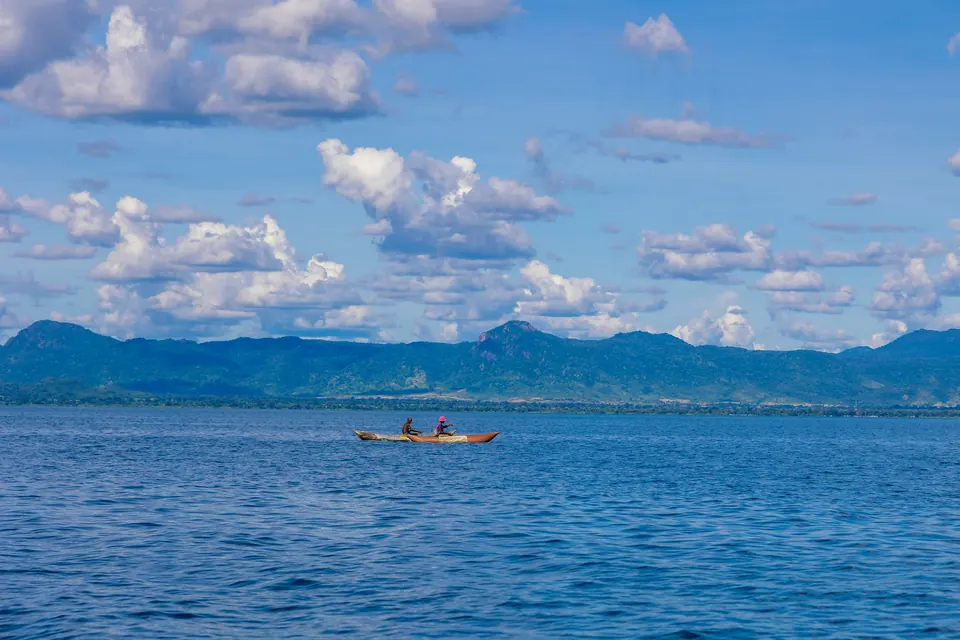 Two people in a small boat on the calm blue water of Lake Malawi, with hills beyond.
