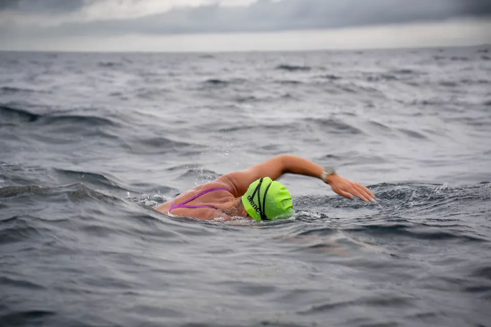 A swimmer in open water under grey skies, seen from the escort boat.