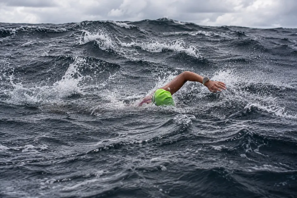 A marathon swimmer in a green cap swimming through rough, white-capped seas.